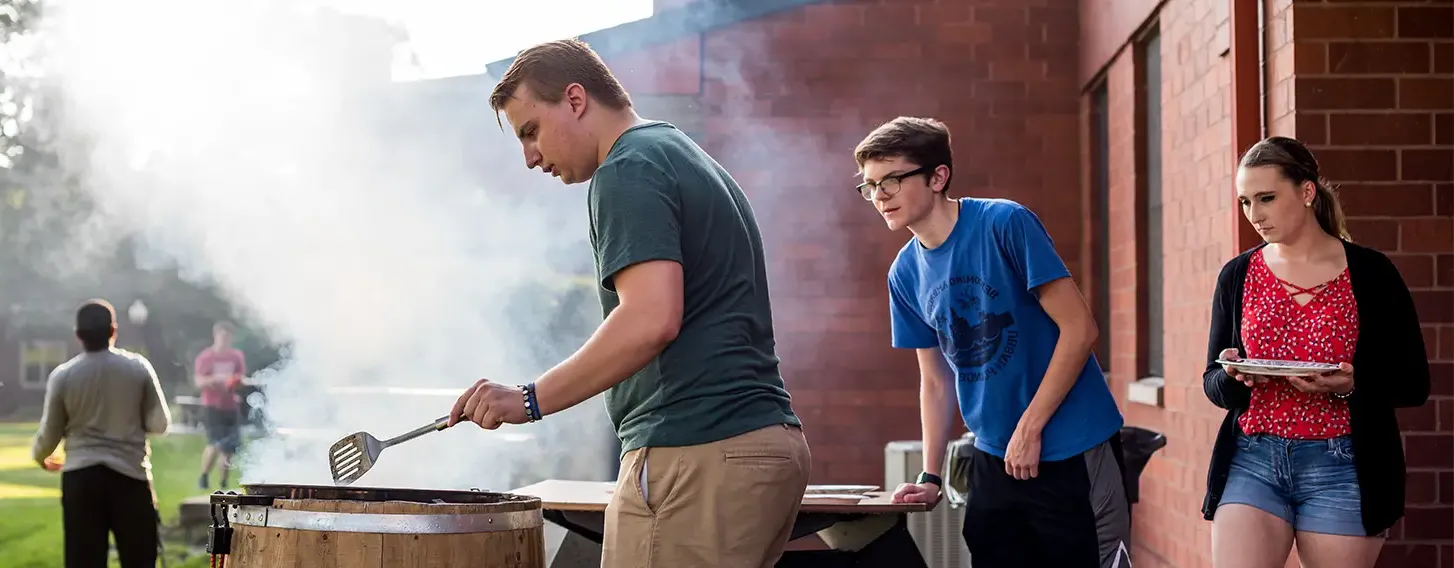 A gathering of students surrounding a barrel smoking food at upperclassmen housing.